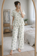 Woman wearing Blush Whispers cream peplum night suit with blue floral prints and full-length pants, taking a mirror selfie in a cozy bedroom.